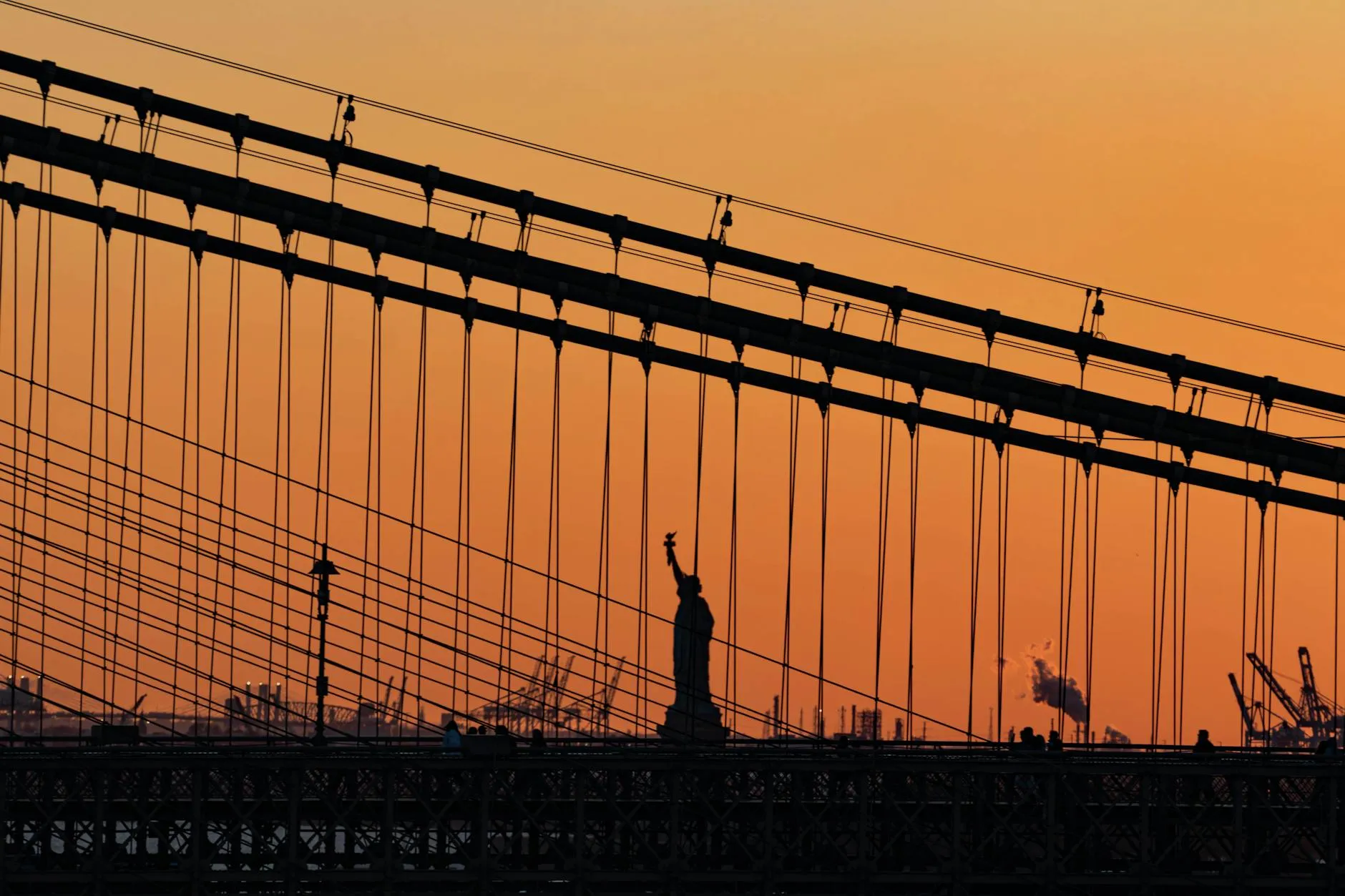 Silhouette of the Statue of Liberty against a sunset sky framed by bridge cables