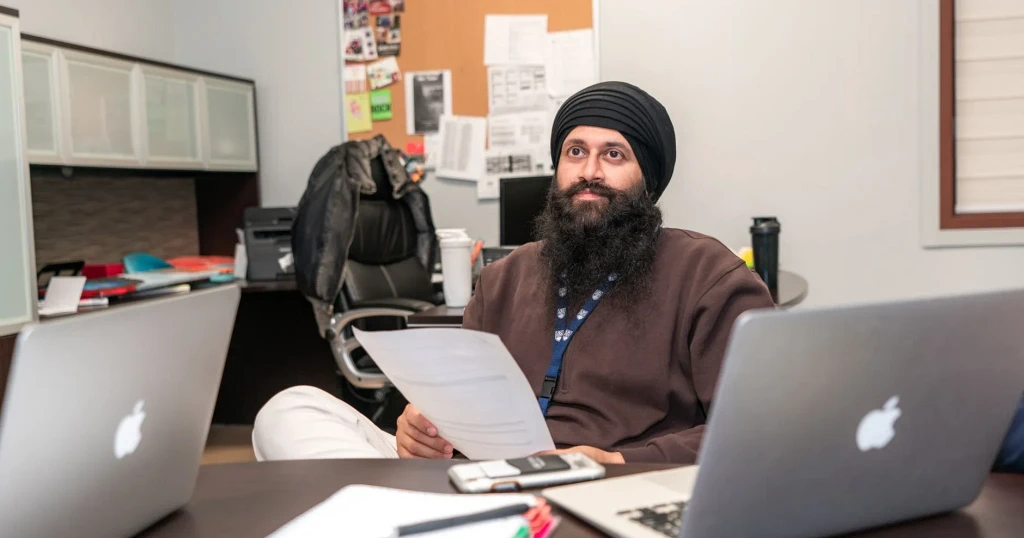 Hombre con turbante sentado en un escritorio de oficina entre dos computadoras portátiles, leyendo un documento impreso