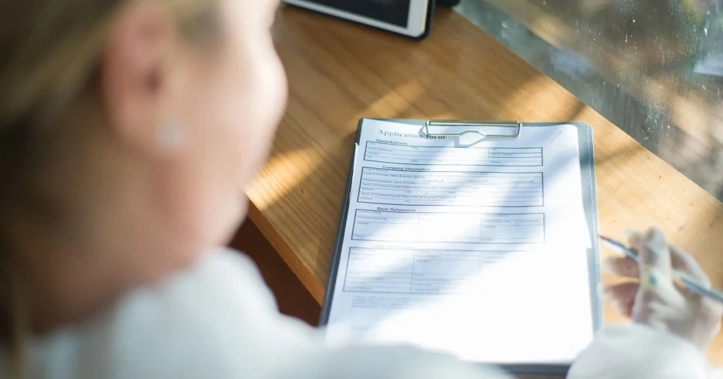 Mujer sentada junto a una ventana revisando un formulario de solicitud en un portapapeles