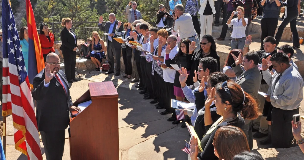 Group of new citizens raising their right hands to take the oath outdoors, facing an official at a podium beside flags