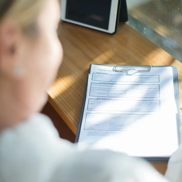 Mujer sentada junto a una ventana revisando un formulario de solicitud en un portapapeles