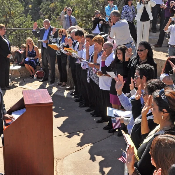Group of new citizens raising their right hands to take the oath outdoors, facing an official at a podium beside flags